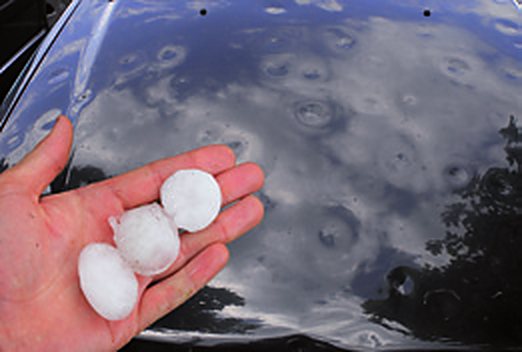 Hagelkörner in einer Hand vor einer von Hagel verbeulten Fahrzeugfläche
